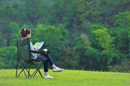 young woman sat on camping stool in the greenery looking out and reading