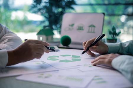 Close-up of two people discussing recycling and circular economy concepts, with a recycling symbol on documents and sustainability-related icons displayed on a laptop in the background.