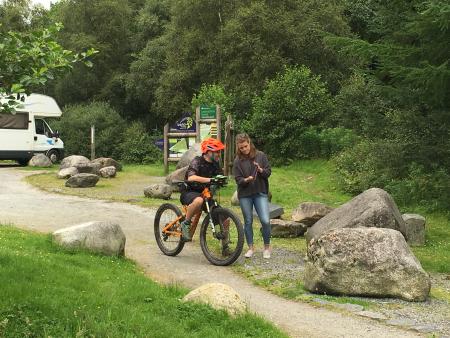 A mountain biker in a rural car park looking at a tablet screen which is held by another person.