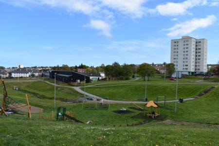 A sunken area of greenspace with a play area, there are paths running down to the play area and tower blocks behind it.