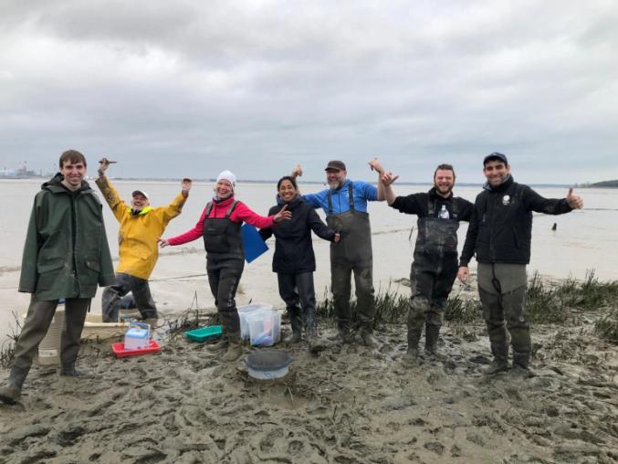 A group of people on a muddy shoreline smiling, they are wearing waders and are muddy.
