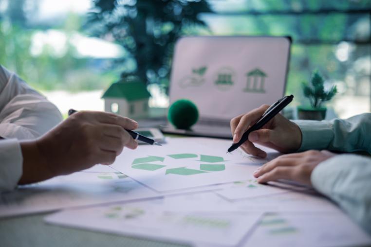 Close-up of two people discussing recycling and circular economy concepts, with a recycling symbol on documents and sustainability-related icons displayed on a laptop in the background.