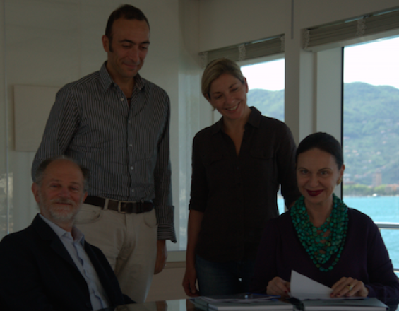 Four people, two sitting and two standing behind them in a room with a view over a body of water.