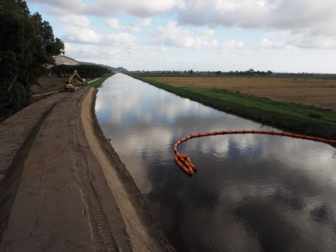 A river with a digger working on the left bank, agricultural fields on the right bank and red floating tubing on the water surfave
