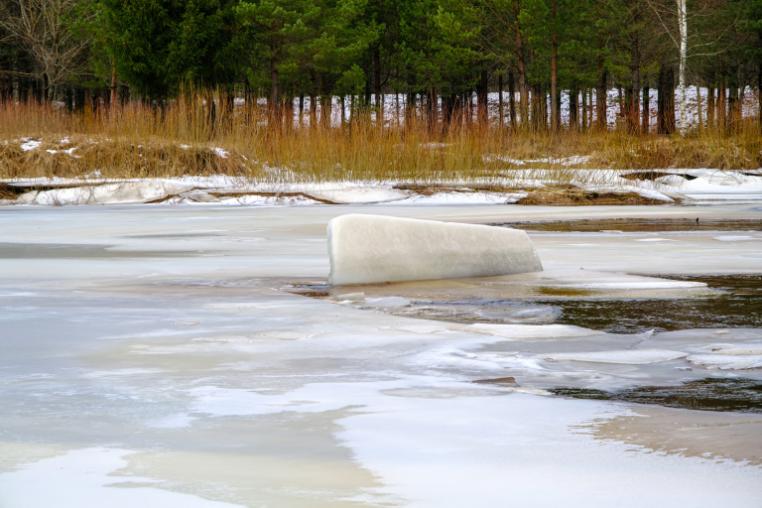 Large lump of ice on a river covered with broken ice, there are some trees in the background on the shore.