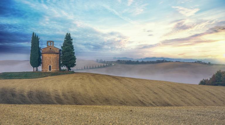 Carapello chapel with a tall tree either side of the building in rolling agricultural land, there is mist over the fields. 