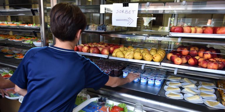 A child reaching for a yoghurt from metal shelves stocked with fruit, vegetables and dairy products.