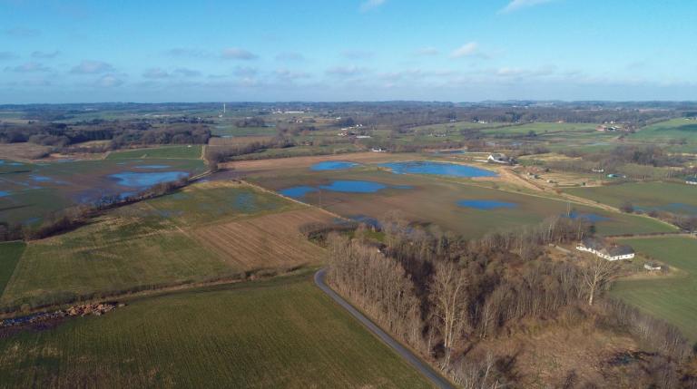 Elevated view of expansive farmland with patches of woodland, some fields have very large puddles.