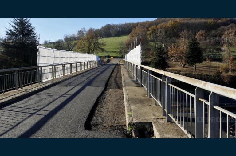 A bridge over a road with metal railings, the bridge leads to an open field on a slope with significant woodland fringing it.